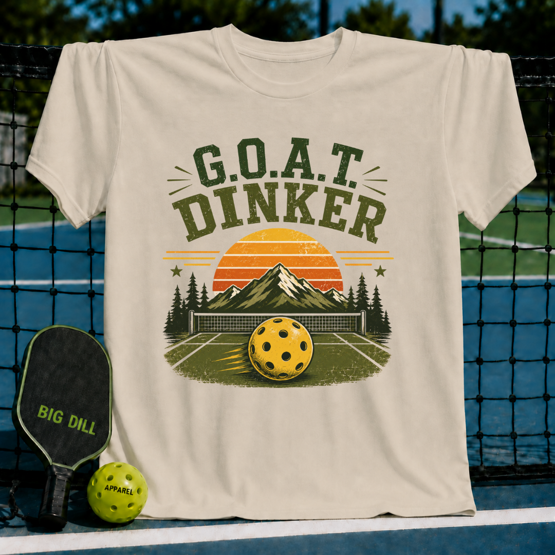 Beige unisex t-shirt featuring a retro pickleball graphic with a sunset, mountains, and G.O.A.T. DINKER text on a court background.