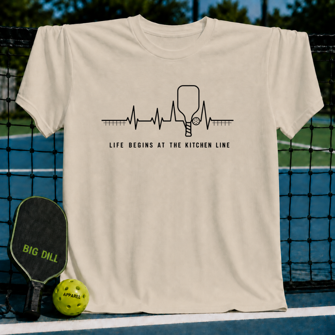Beige unisex t-shirt featuring a black heartbeat graphic with a pickleball paddle and ball, text reads Life Begins At The Kitchen Line.