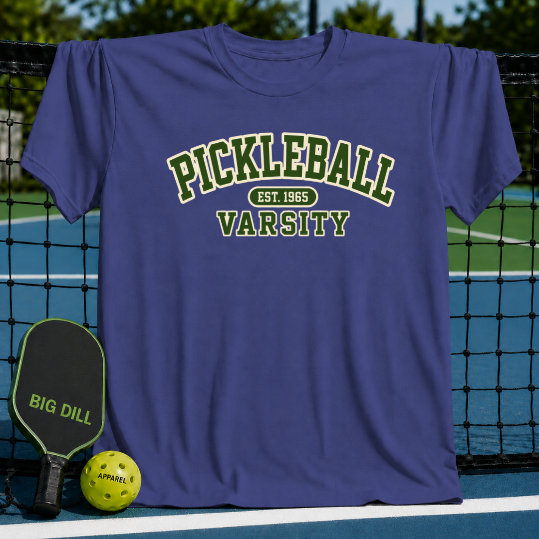 Blue crewneck t-shirt with green and white Pickleball Varsity text, displayed on a pickleball court with a paddle and ball.