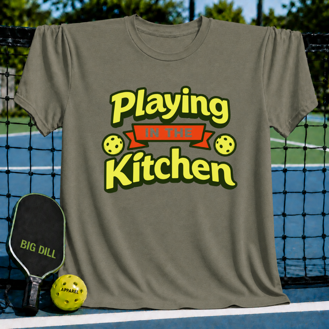 Olive green crewneck t-shirt featuring a yellow Playing in the Kitchen pickleball graphic on a court background.