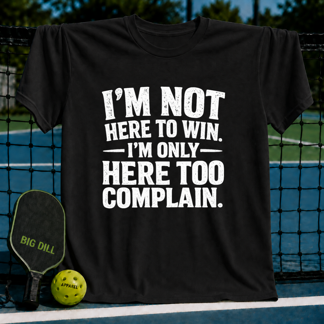 Black unisex t-shirt with distressed white text reading I'm Not Here To Win, I'm Only Here To Complain, displayed on a pickleball court.