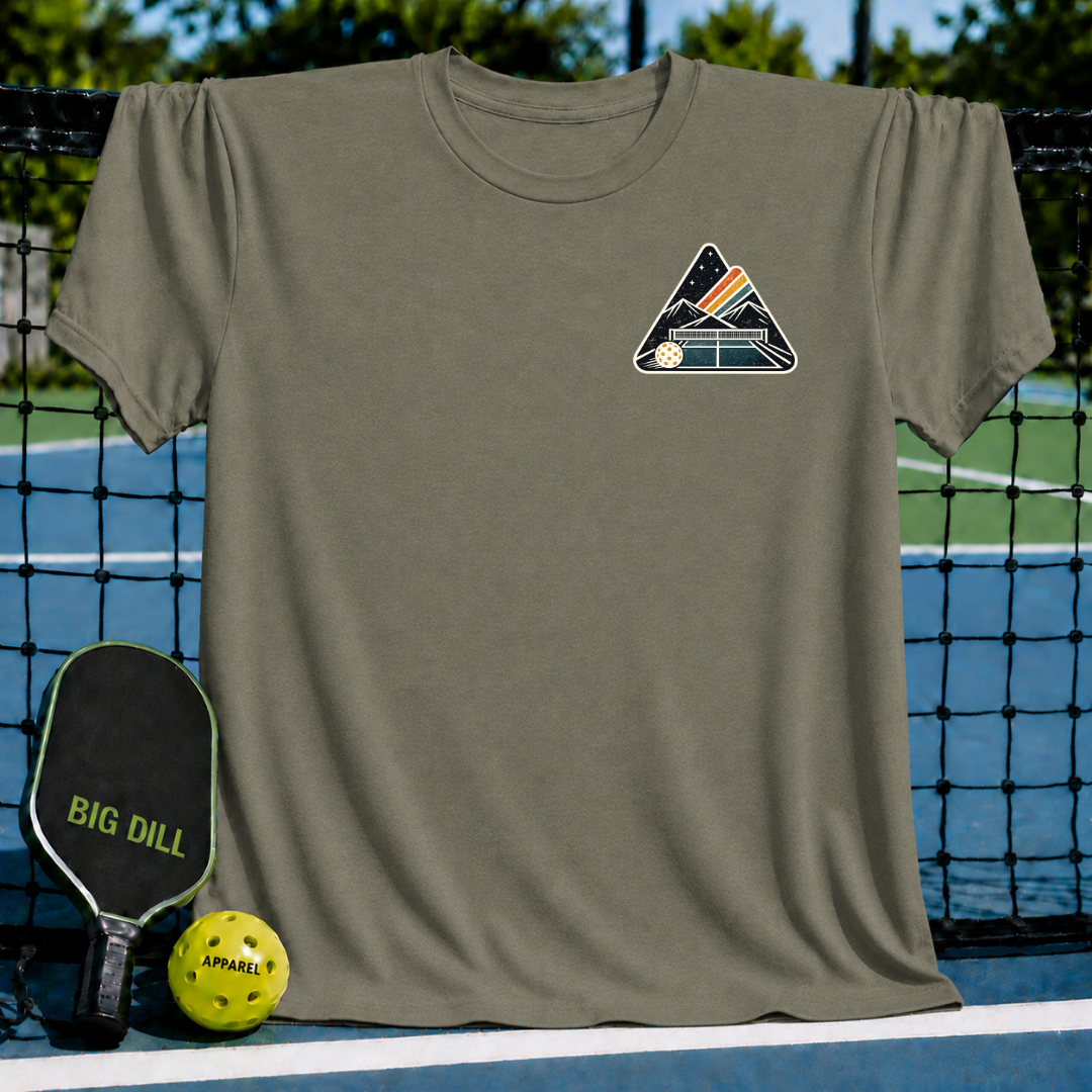 Olive green unisex t-shirt featuring a mountain and pickleball court crest graphic, displayed on a pickleball court net.