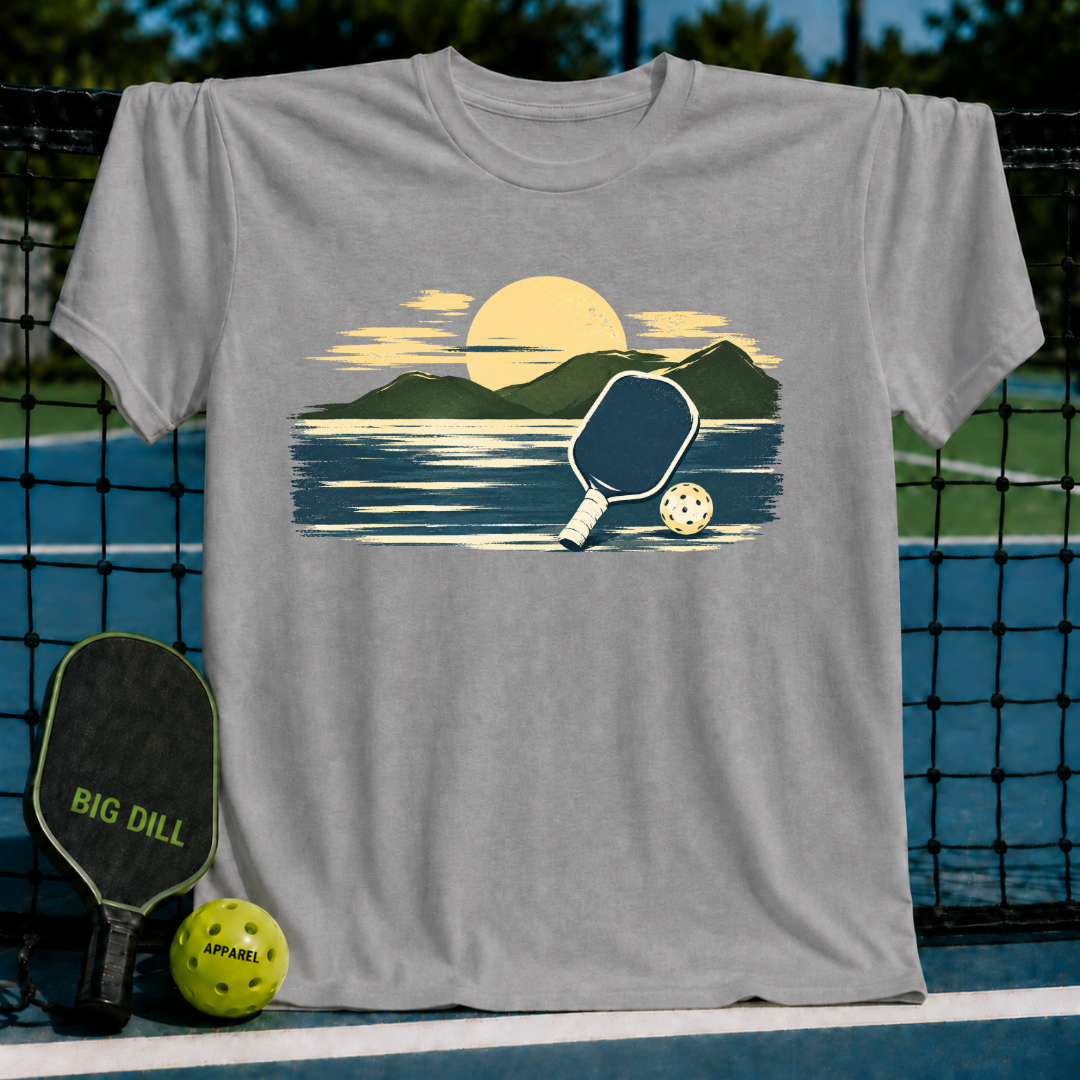 Grey unisex t-shirt featuring a painterly mountain and pickleball graphic, displayed on a court with a paddle and ball.