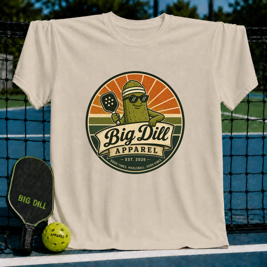 Beige Big Dill Apparel t-shirt featuring a retro pickleball mascot graphic with a pickle in sunglasses and a visor.