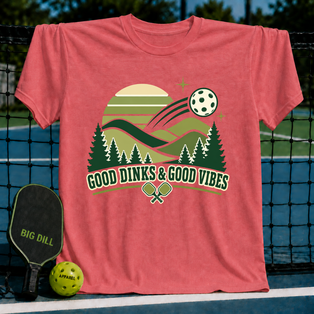 Coral pink t-shirt featuring a retro pickleball landscape graphic with pine trees, a sun, and Good Dinks & Good Vibes text.