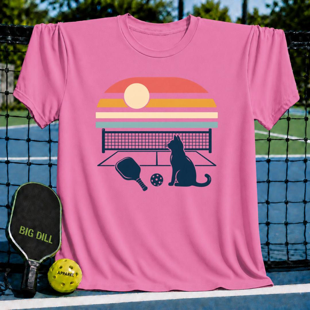 Pink t-shirt featuring a retro sunset, a cat, and a pickleball court graphic hanging on a court net.