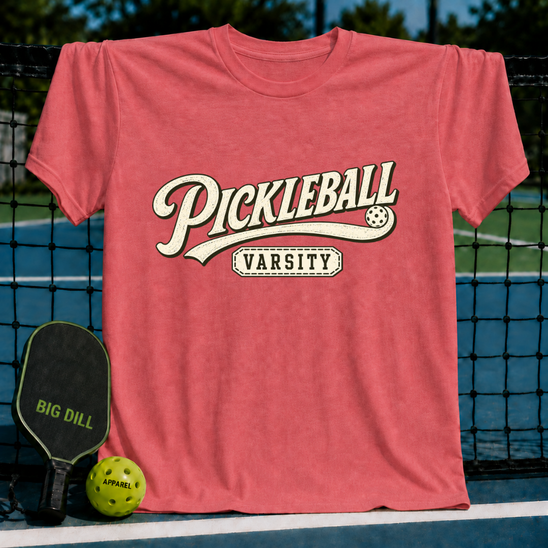 Coral red crewneck t-shirt featuring vintage-style Pickleball Varsity graphic lettering on a pickleball court background.