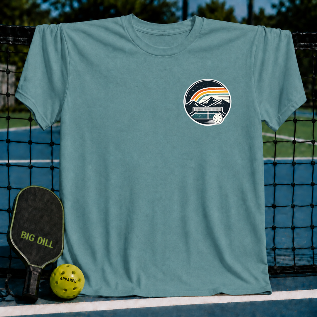 Blue-grey unisex t-shirt featuring a circular pickleball court graphic with mountains and a rainbow, displayed on a court.