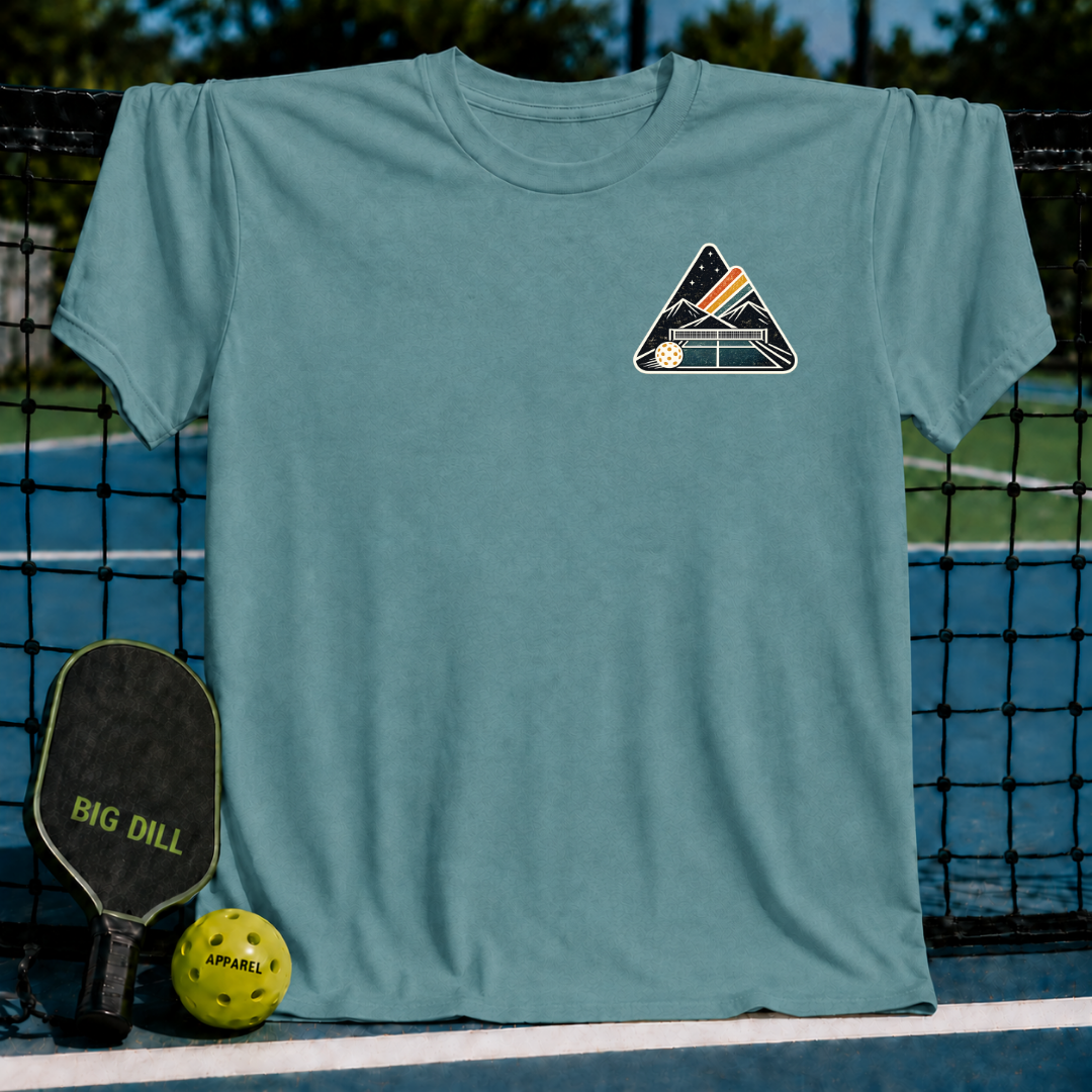 Blue-grey unisex t-shirt featuring a mountain and pickleball court crest graphic, displayed on a pickleball court.