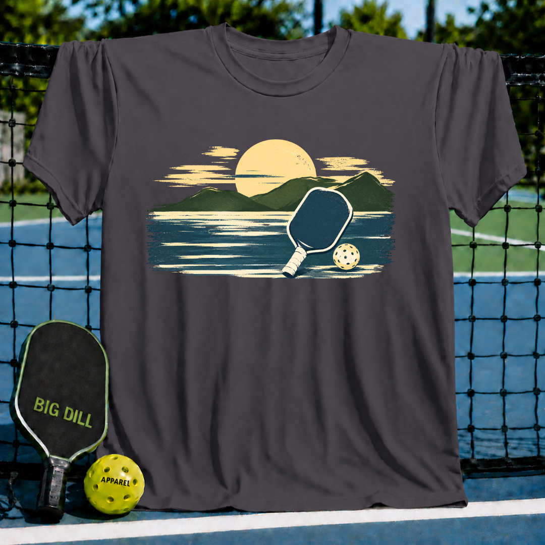 Olive green unisex t-shirt featuring a painterly mountain and pickleball graphic in golden and navy tones.