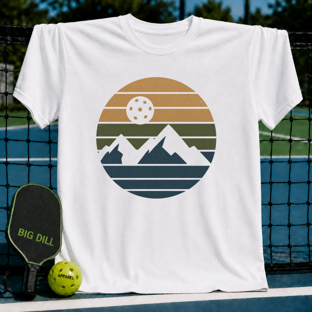 White unisex t-shirt featuring a retro circular graphic of mountain peaks and a pickleball sun in tan, olive, and teal tones.