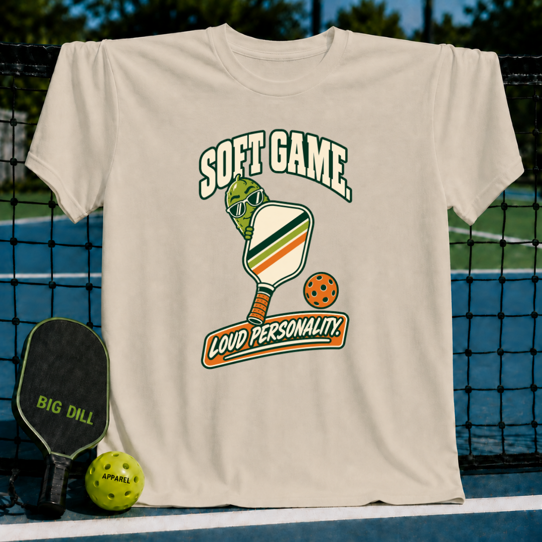 Beige cotton t-shirt featuring a pickleball-themed graphic with a cartoon pickle, paddle, and Soft Game Loud Personality text.