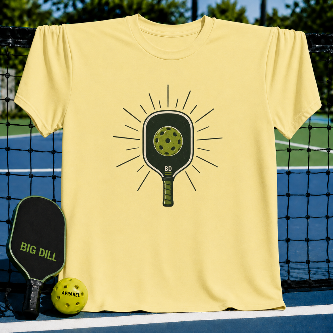 Light yellow crew neck t-shirt featuring a graphic of a pickleball paddle and ball on a pickleball court background.