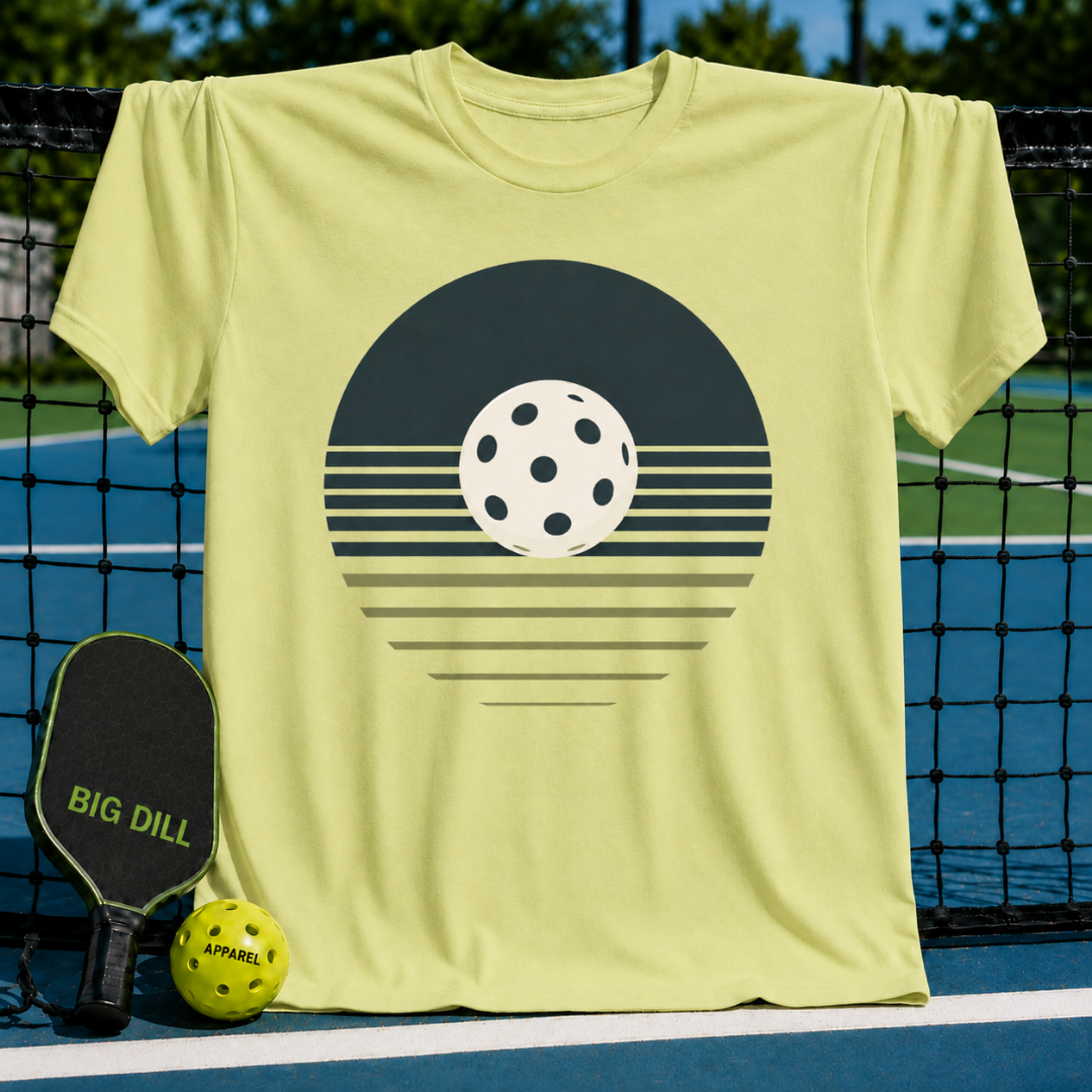 Light yellow unisex t-shirt featuring a retro charcoal pickleball sunset graphic, displayed on a pickleball court.