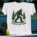 White unisex t-shirt featuring a green Sasquatch illustration holding a pickleball paddle with the text Elusive Soft Game.