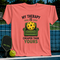 Coral unisex t-shirt featuring a graphic of a yellow pickleball sitting in a green armchair with the text My Therapy Is Cheaper.