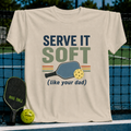 Cream unisex t-shirt featuring retro Serve It Soft pickleball typography, a paddle, and a ball graphic on a court background.