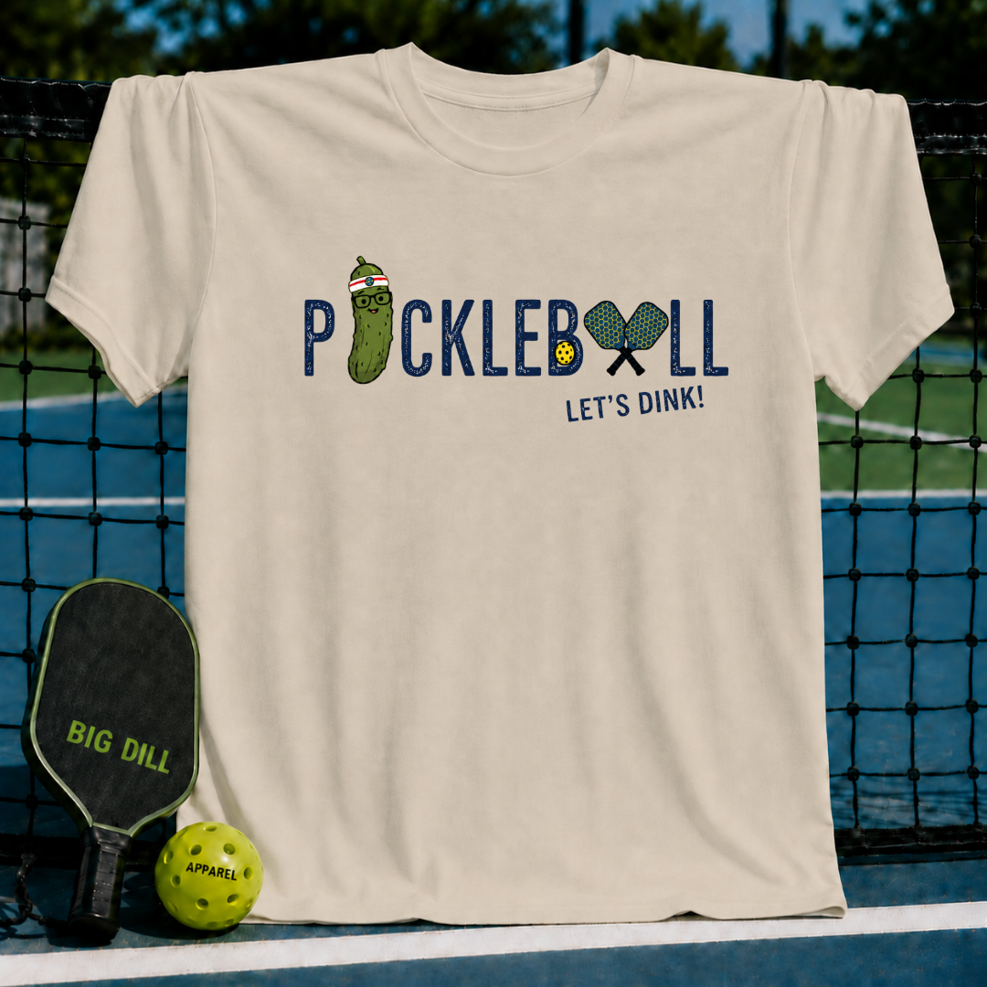 Beige crewneck t-shirt featuring a Pickleball Let's Dink graphic with a pickle character and paddles on a pickleball court.