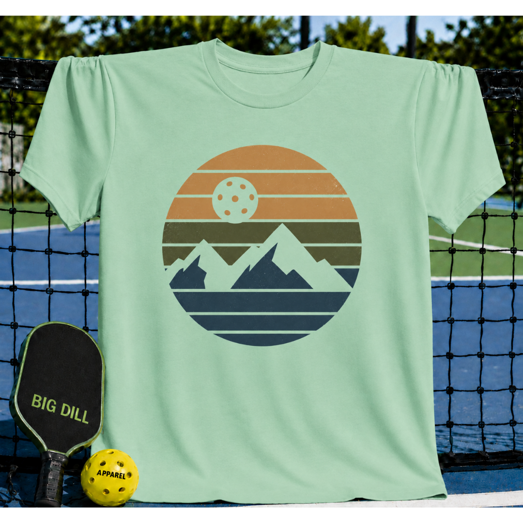 Mint green unisex t-shirt featuring a retro circular mountain graphic with a pickleball sun, displayed on a court.