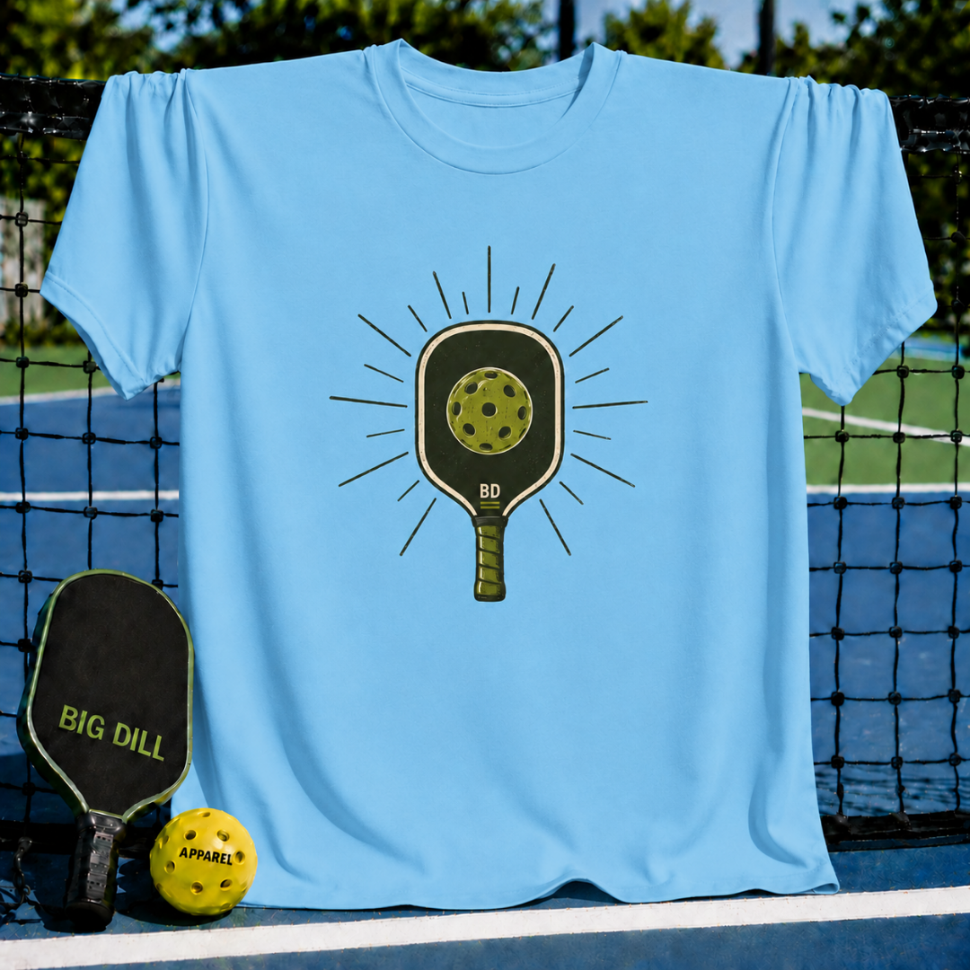 Light blue crew neck t-shirt featuring a vintage-style pickleball paddle and ball graphic on a pickleball court background.