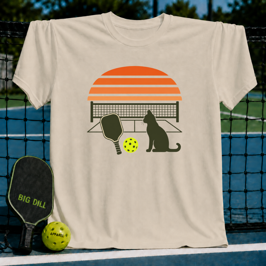 Light beige unisex t-shirt featuring a retro sunset, cat silhouette, pickleball paddle, and ball graphic.