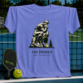 Periwinkle blue unisex T-shirt featuring a graphic of The Thinker holding a pickleball paddle with a neon yellow ball.