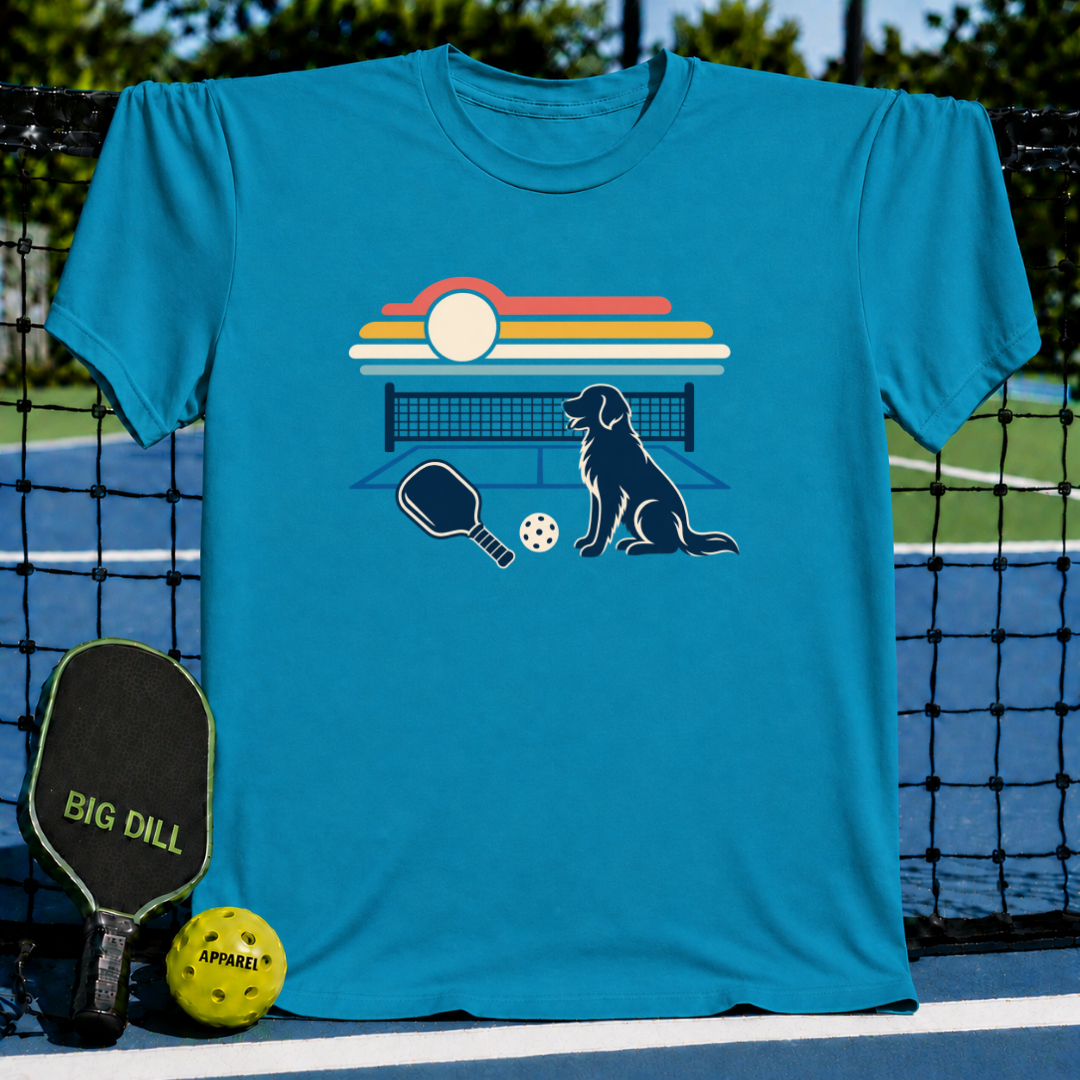 Bright blue t-shirt featuring a retro sunset, pickleball court, and dog graphic, displayed on a pickleball court background.
