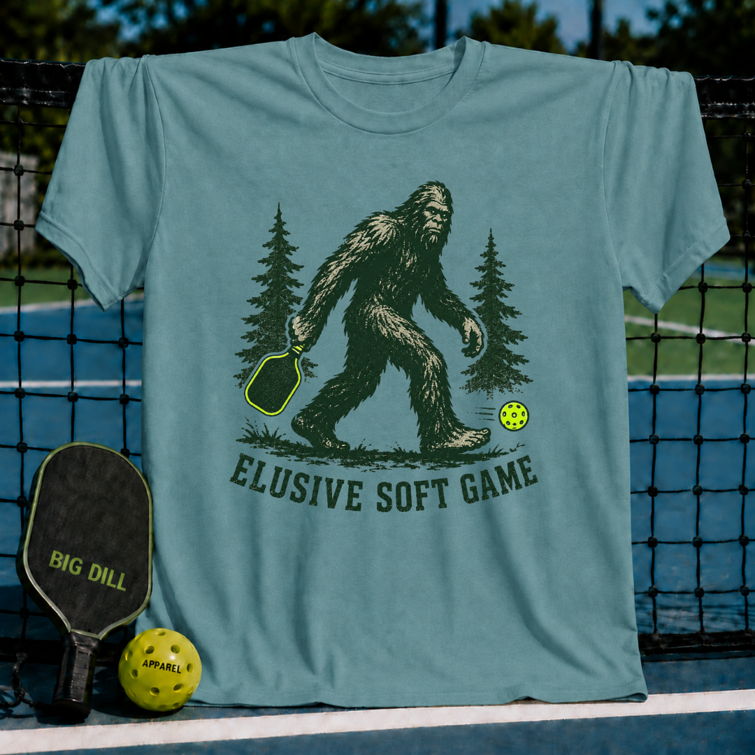 Blue-grey crewneck t-shirt featuring a green Sasquatch graphic with a pickleball paddle and neon ball, labeled Elusive Soft Game.