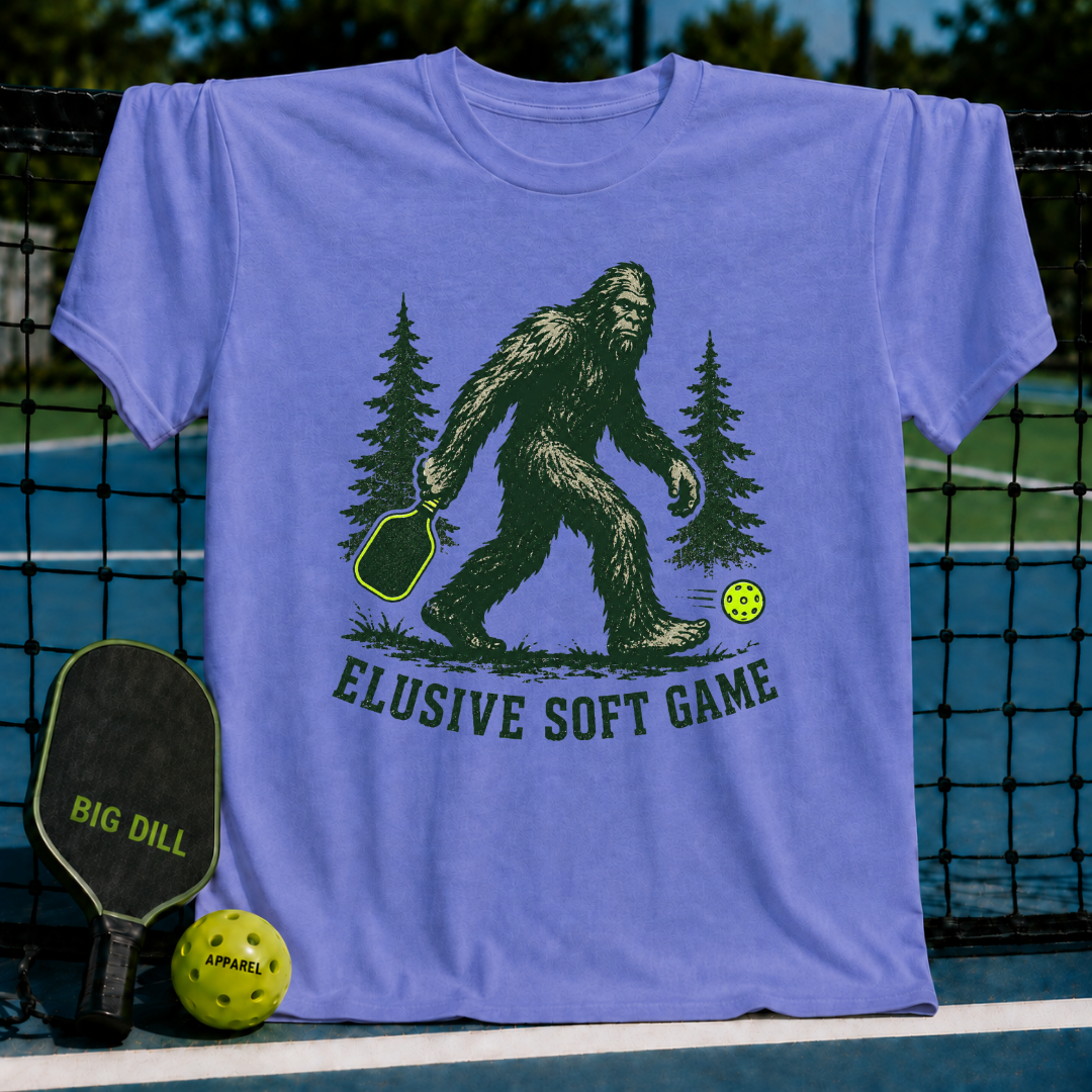 Periwinkle t-shirt featuring a green Sasquatch graphic with a pickleball paddle and neon ball, text reading Elusive Soft Game.