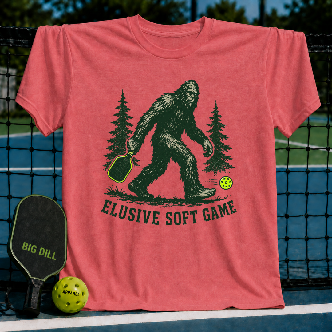Coral unisex t-shirt featuring a green Bigfoot illustration, pickleball paddle, and the text Elusive Soft Game.