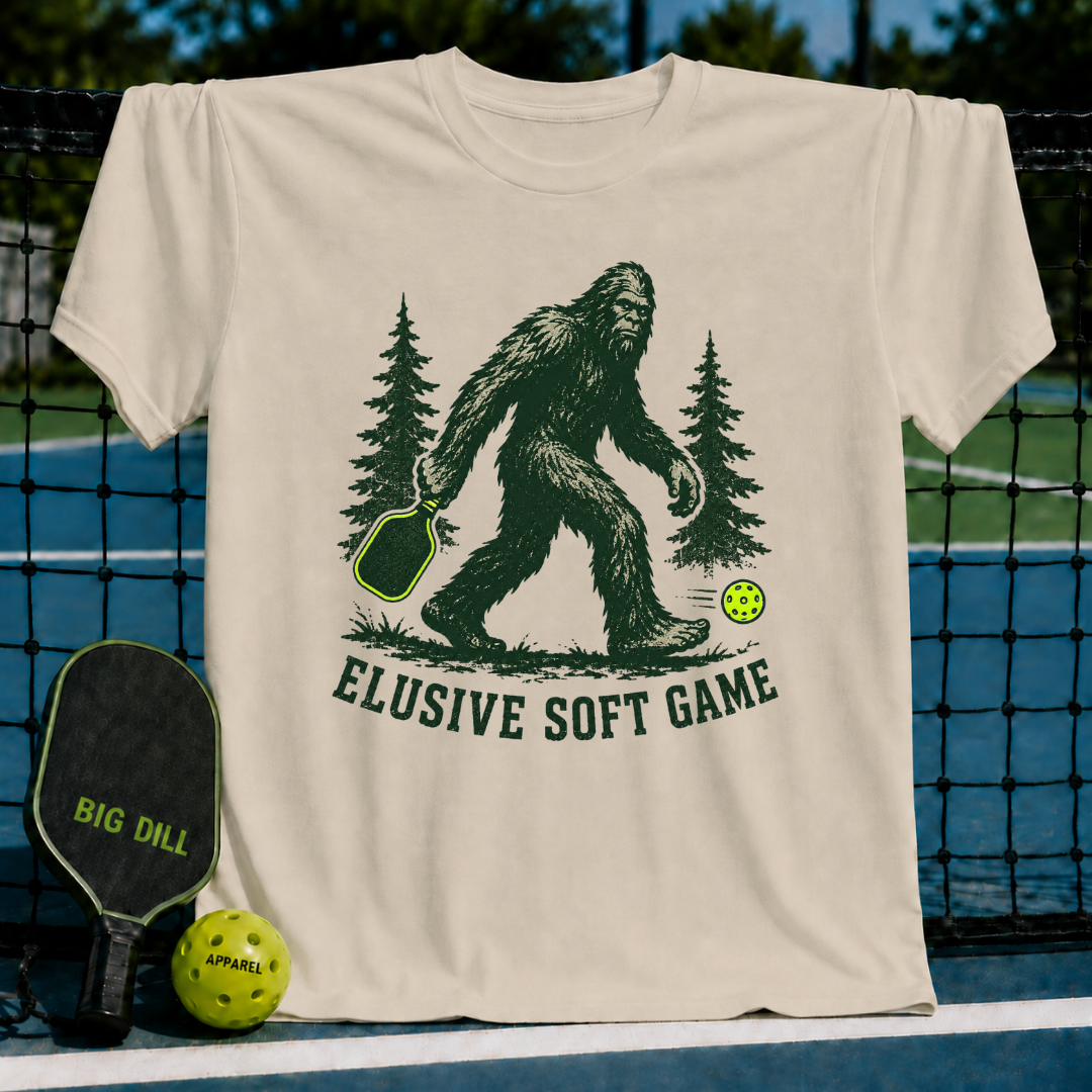 Cream-colored unisex t-shirt featuring a green Sasquatch illustration with a pickleball paddle and neon ball, labeled Elusive Soft Game.