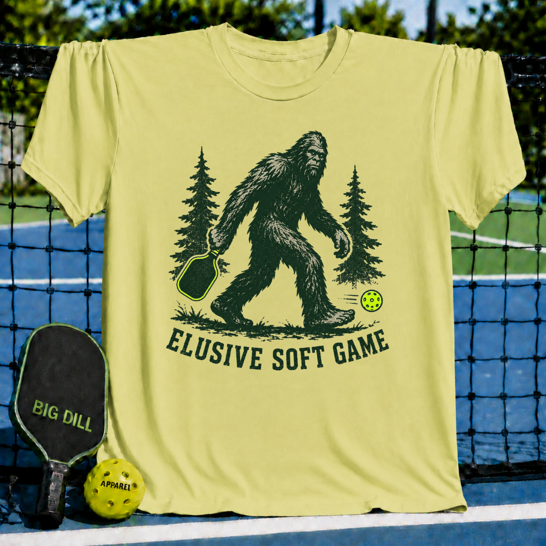 Light yellow unisex t-shirt featuring a green Sasquatch graphic with a pickleball paddle and neon ball, text reads Elusive Soft Game.