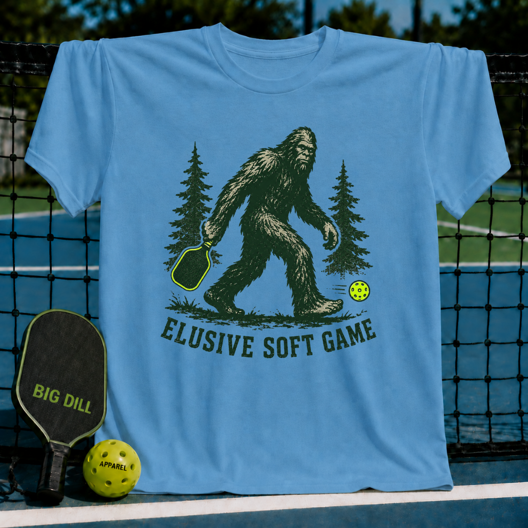 Light blue unisex t-shirt featuring a green Sasquatch graphic with a pickleball paddle and neon ball, labeled Elusive Soft Game.