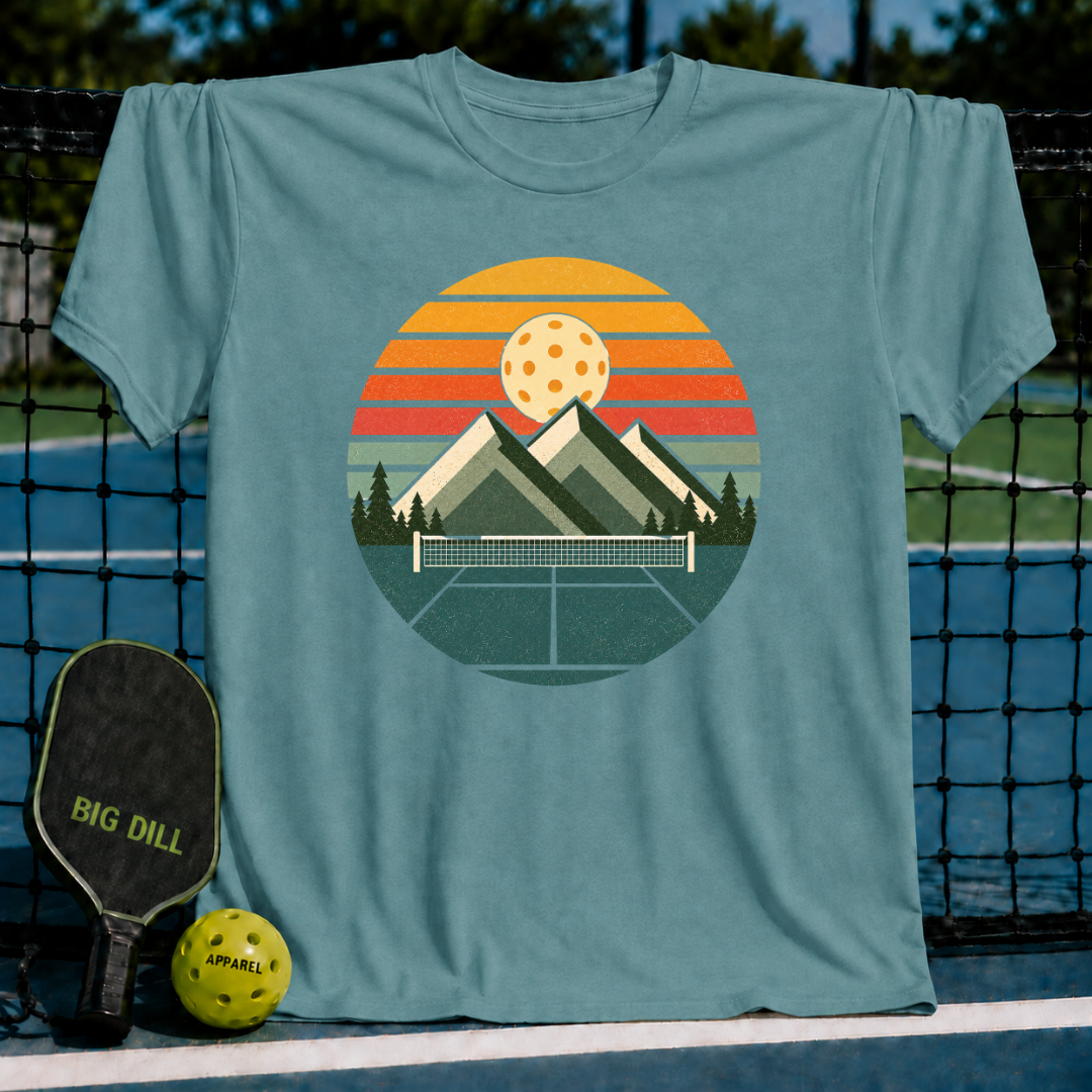 Deep teal unisex t-shirt featuring a retro circular graphic of mountains, a pickleball court, and a sunset-striped sky.
