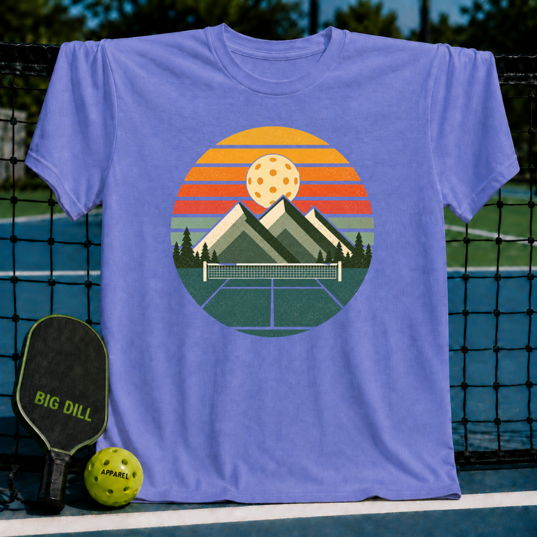 Periwinkle blue unisex t-shirt featuring a retro circular graphic of mountains, a pickleball court, and a sunset.