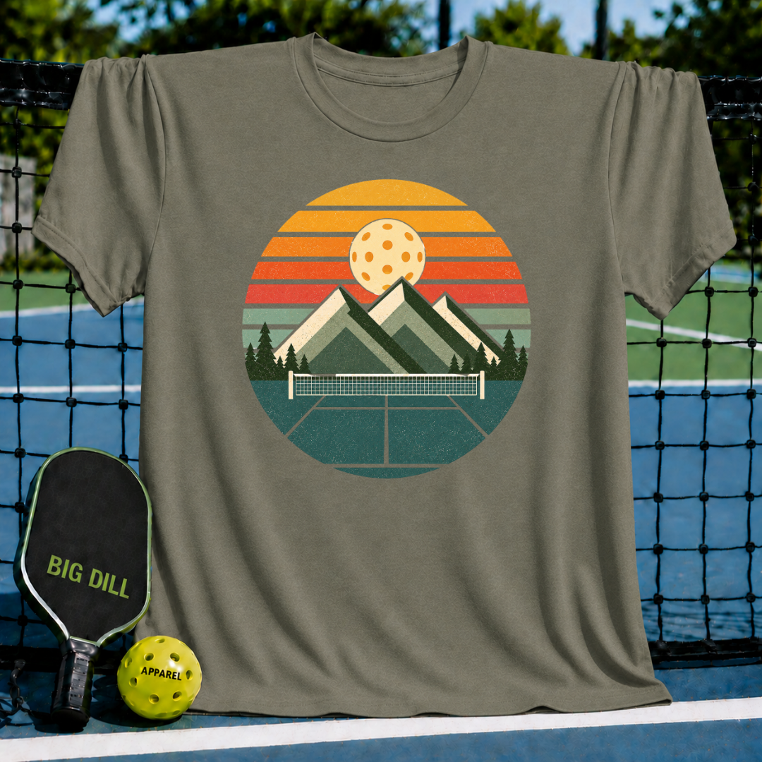 Olive green unisex t-shirt featuring a circular retro mountain, sunset, and pickleball court graphic on a court background.