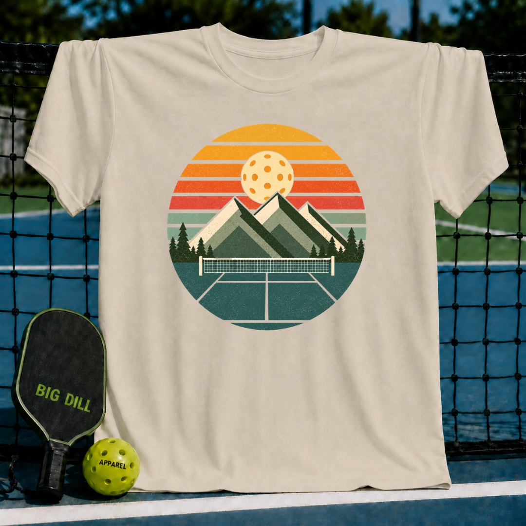 Cream-colored unisex t-shirt featuring a retro circular graphic of mountains, a pickleball net, and a sunset over a court.