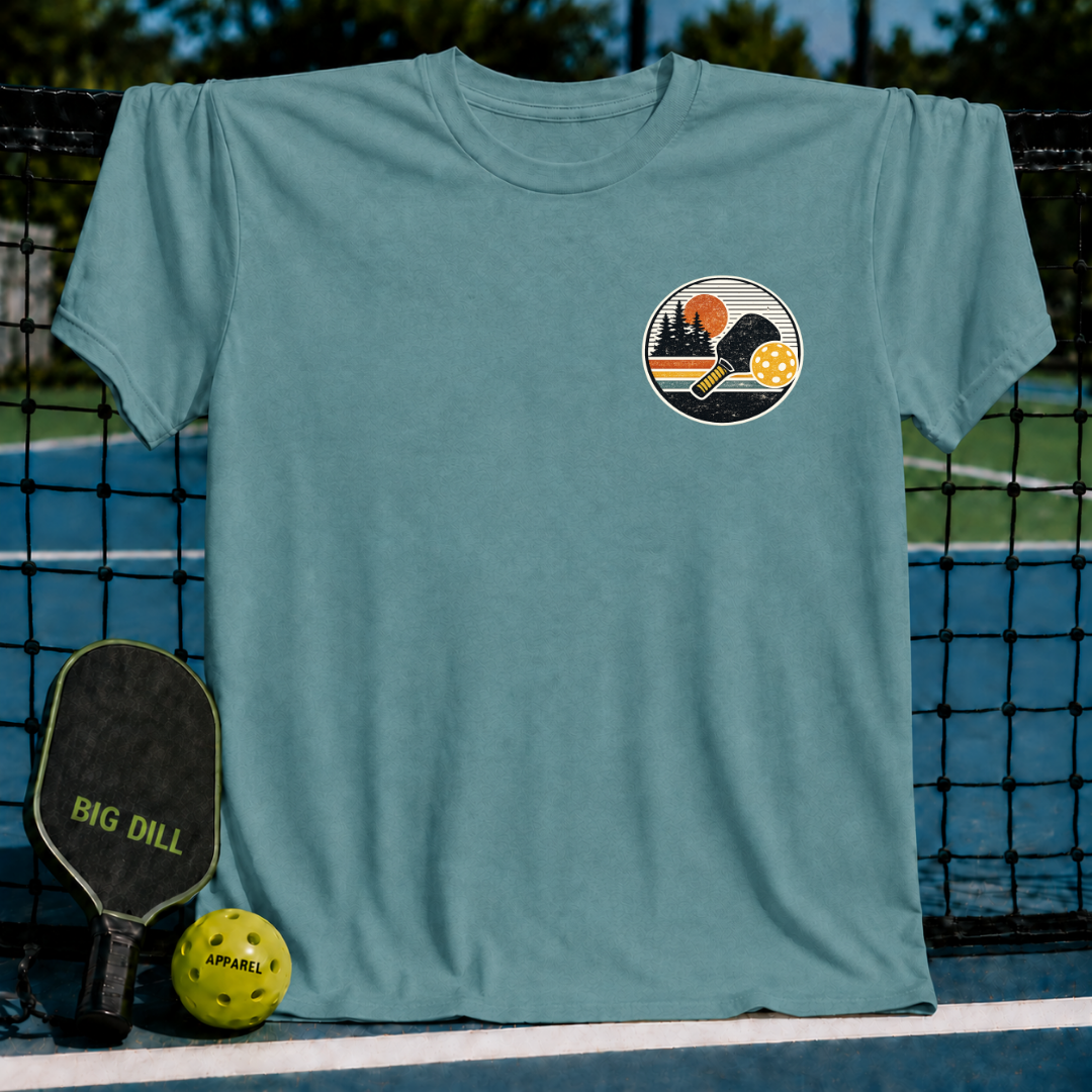 Blue-grey unisex t-shirt featuring a circular sunset pickleball graphic with a paddle and ball on a court background.