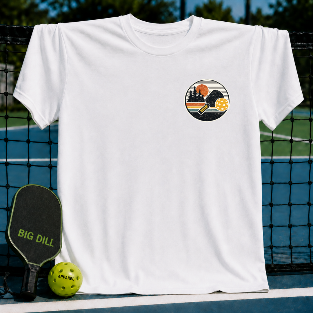White unisex t-shirt featuring a circular sunset crest graphic with a pickleball paddle and ball on a pickleball court.