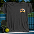 Charcoal grey unisex t-shirt featuring a circular sunset crest graphic with a pickleball paddle and ball on a court.