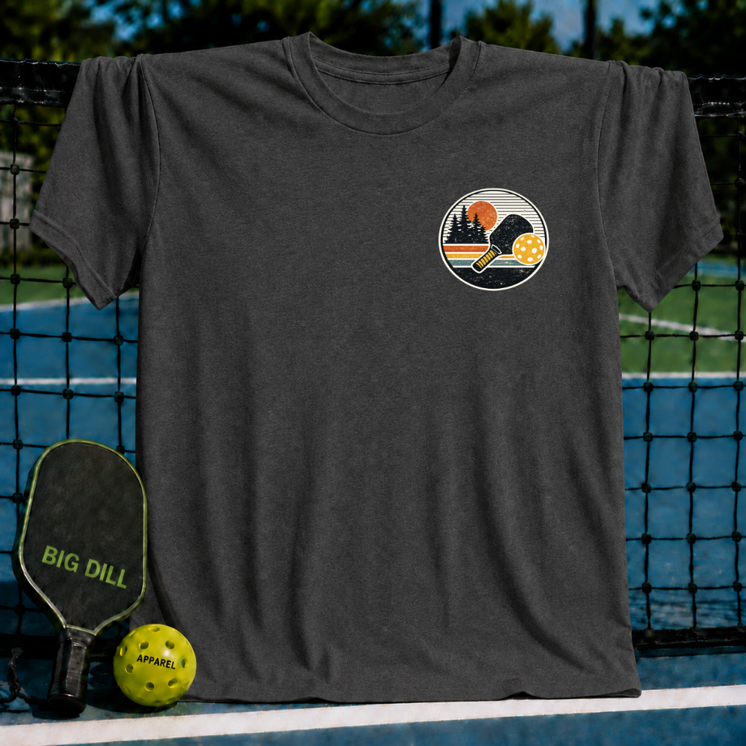 Charcoal grey unisex t-shirt featuring a circular sunset crest graphic with a pickleball paddle and ball on a court.