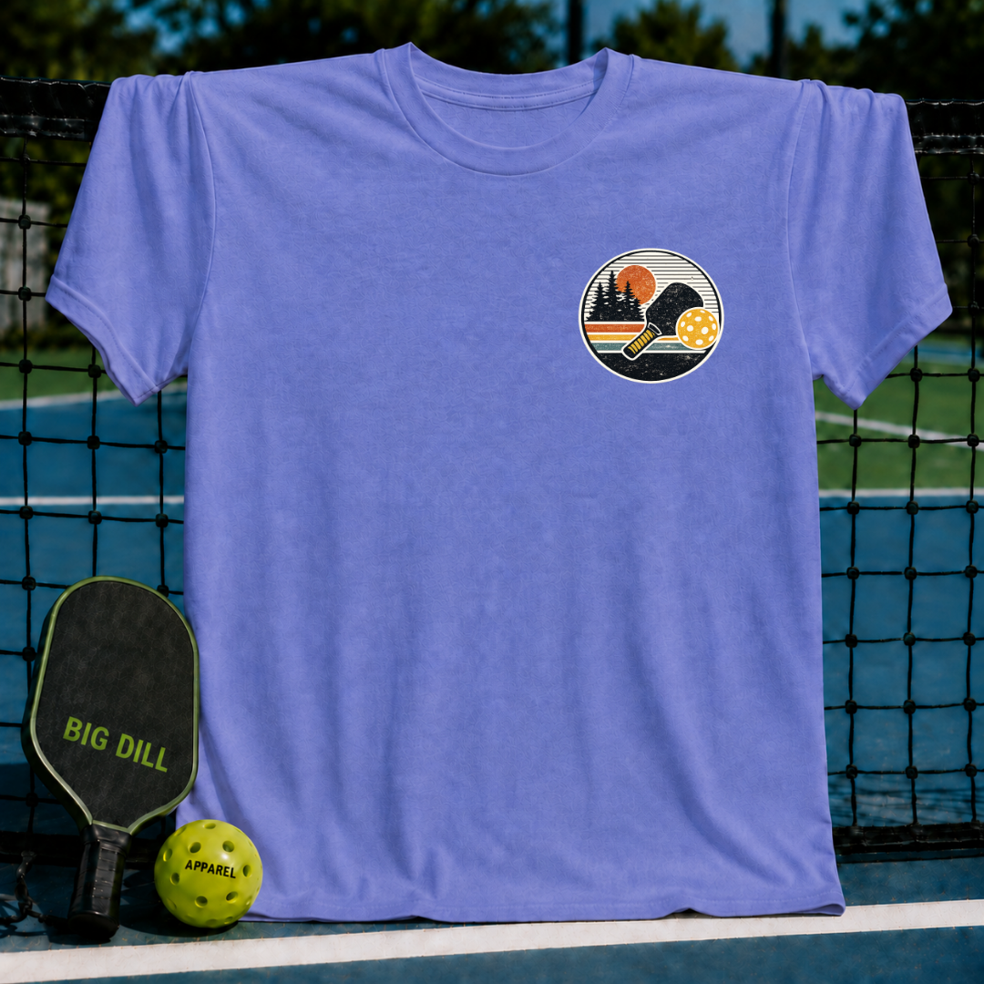 Periwinkle blue unisex t-shirt featuring a circular sunset pickleball graphic with a paddle and ball on a court.