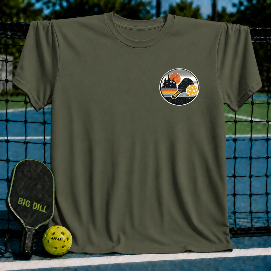 Olive green unisex t-shirt featuring a circular sunset pickleball graphic on the chest, displayed on a court net.