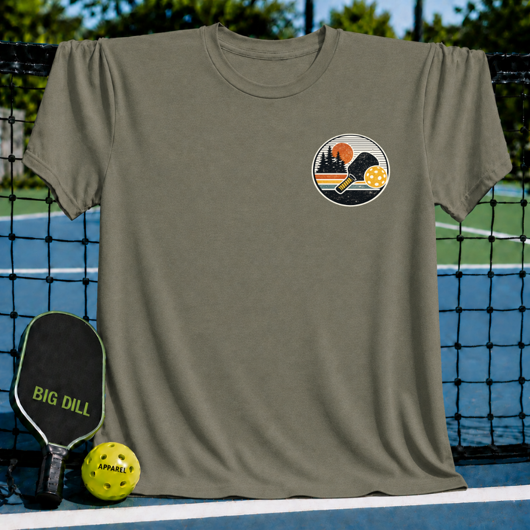 Olive green unisex t-shirt featuring a circular sunset graphic with a pickleball paddle and ball on a pickleball court.