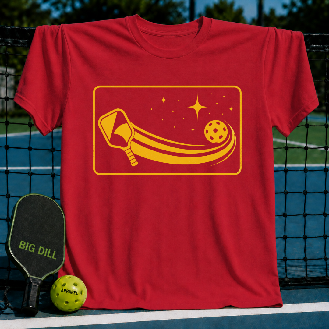 Red unisex t-shirt featuring a yellow cosmic pickleball paddle and ball graphic, displayed on a pickleball court net.
