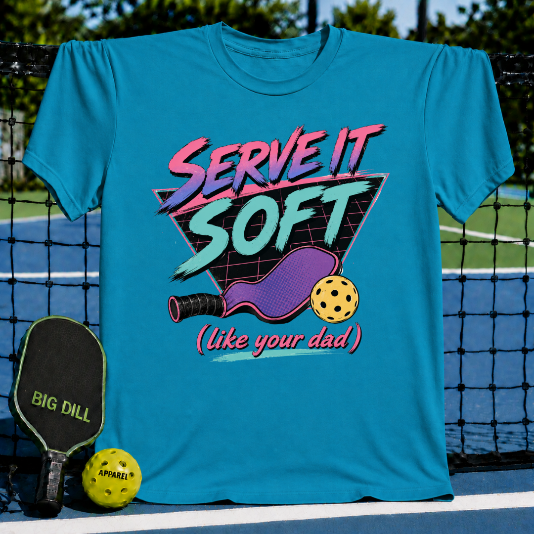 Slate blue unisex t-shirt featuring an 80s neon graphic of a pickleball paddle, ball, and the text Serve It Soft.