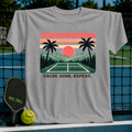 Grey crewneck t-shirt featuring a retro sunset, palm tree, and pickleball court graphic with the text Vacay. Dink. Repeat.