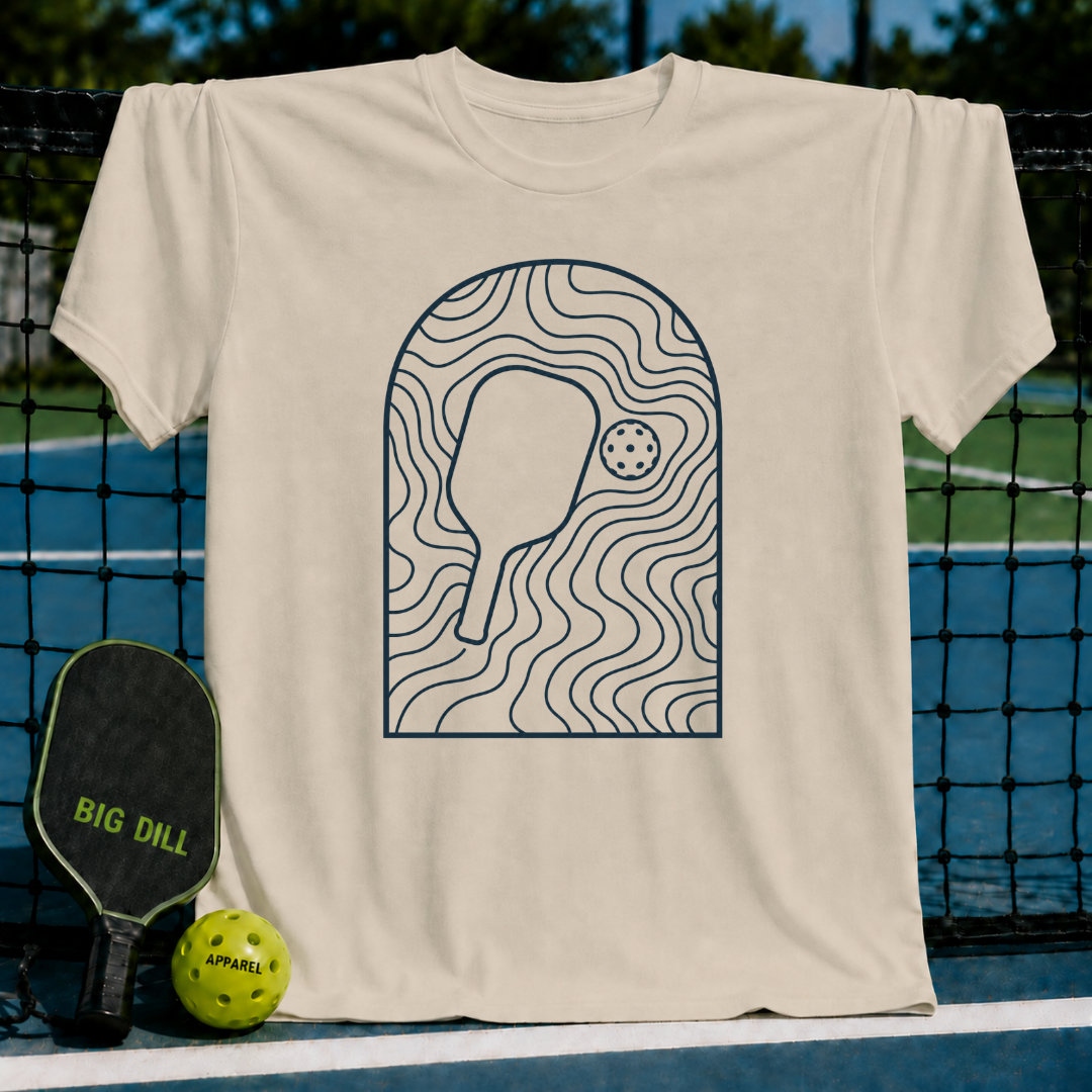 Beige unisex t-shirt featuring a minimalist slate gray arched line art graphic of a pickleball paddle and ball.
