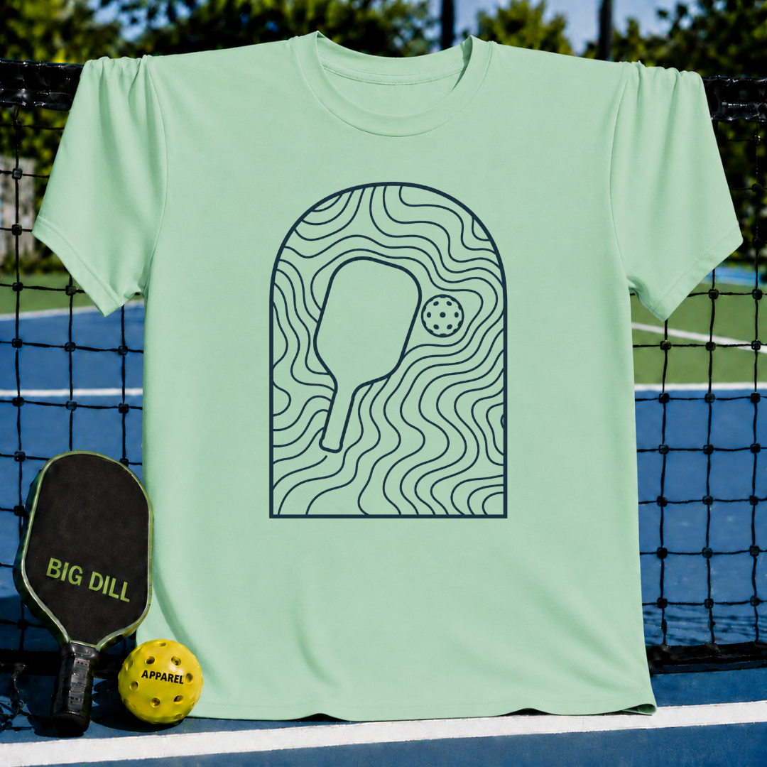 Light green unisex t-shirt featuring a dark gray arched line art graphic of a pickleball paddle and ball.